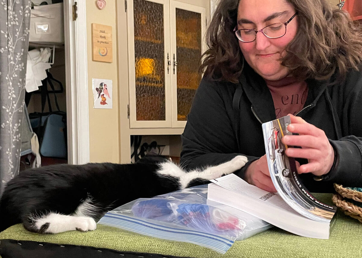 ViK Stohl leaning over reading a book, while cat, Jabbers, while laying down reaches out paw to touch ViK.